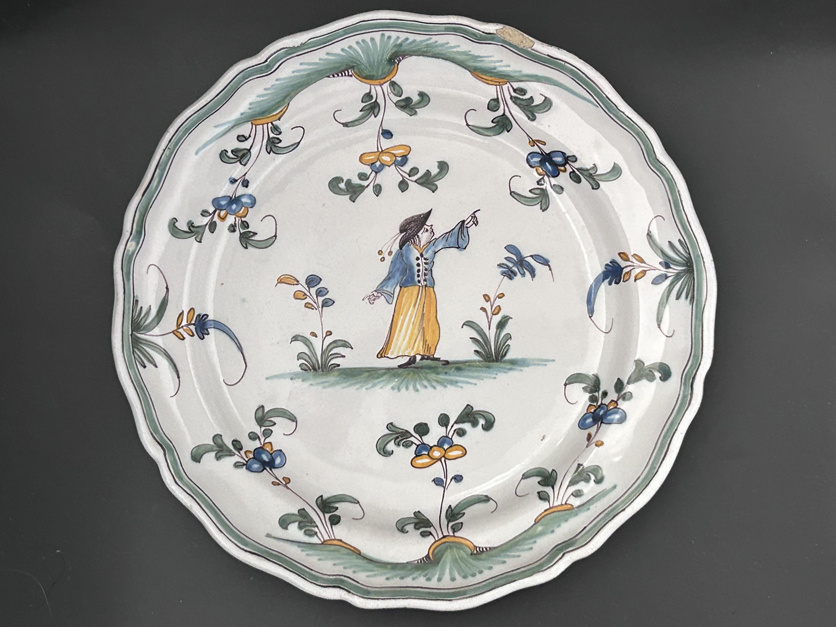 Assiette polychrome de style Moustier, XVIIIe : Assiette en faïence polychromée avec un décor illustrant un personnage indiquant une direction